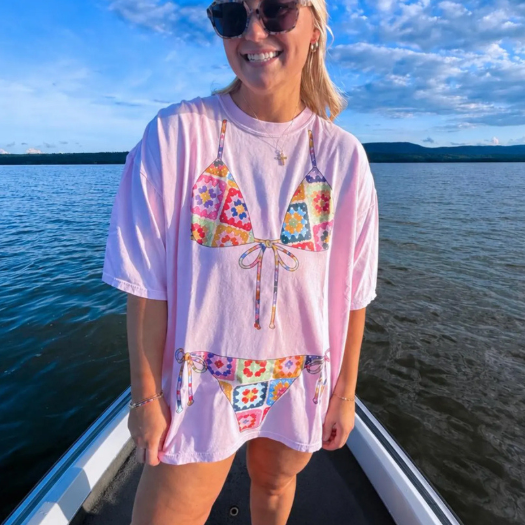 Light pink graphic tee featuring a colorful crochet-style bikini design with patchwork florals and retro summer-inspired artwork.