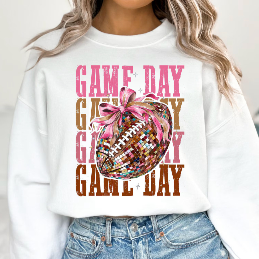 White graphic sweatshirt featuring a disco ball football with a pink bow layered over distressed pink and brown "Game Day" lettering.