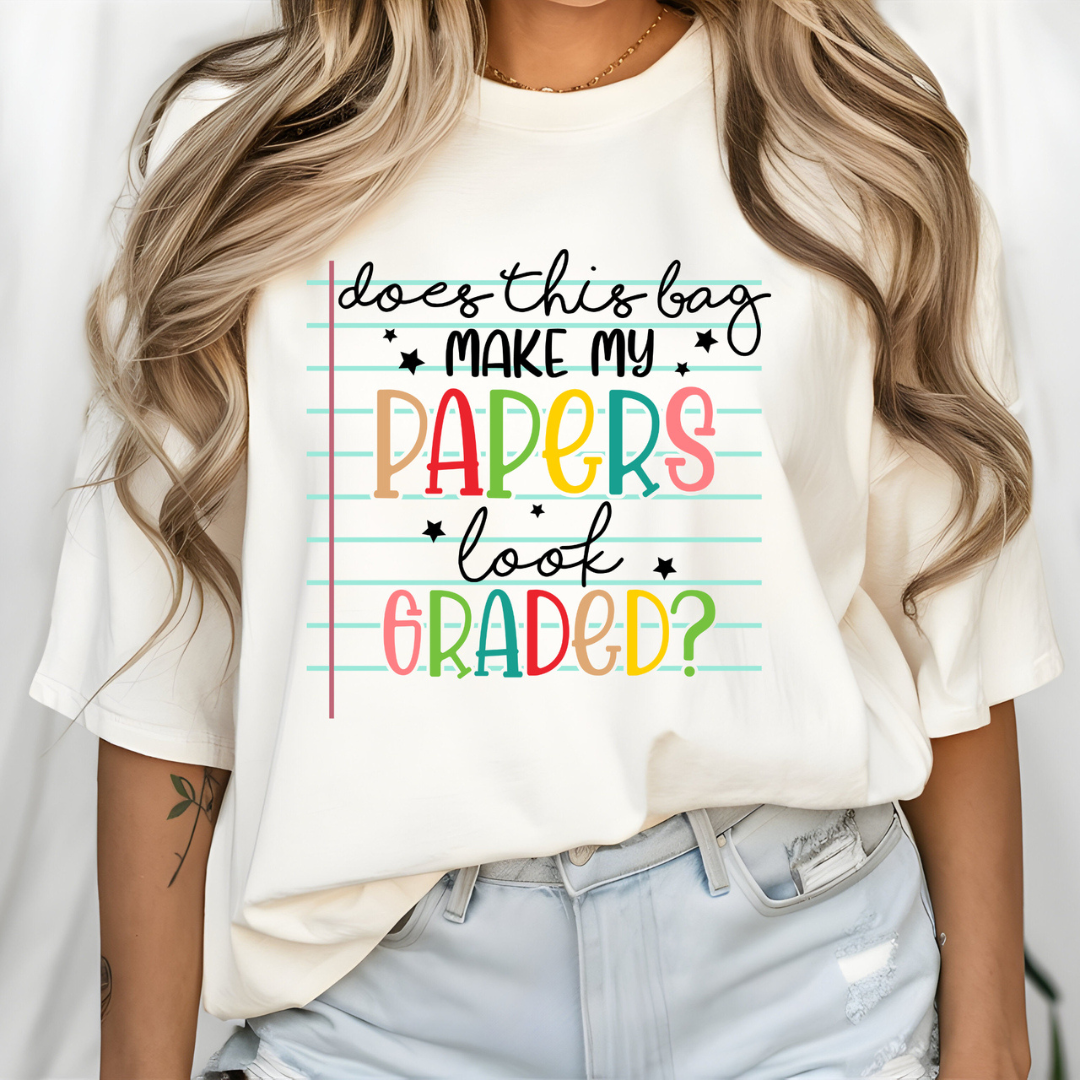 White graphic tee featuring the phrase "Does This Bag Make My Papers Look Graded?" on a colorful notebook paper background.