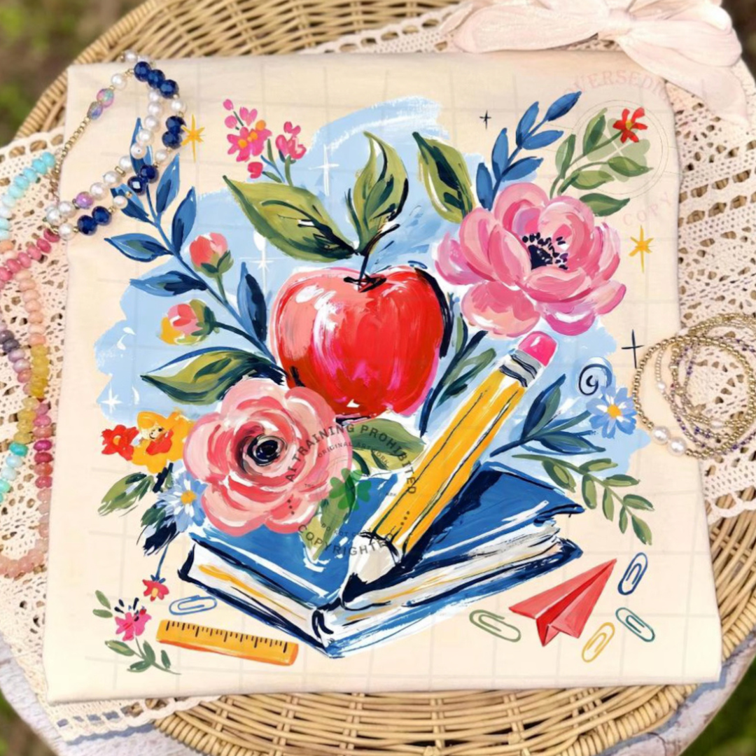 Natural beige graphic tee featuring a watercolor-style teacher design with a red apple, pink flowers, books, pencil, and classroom accents.