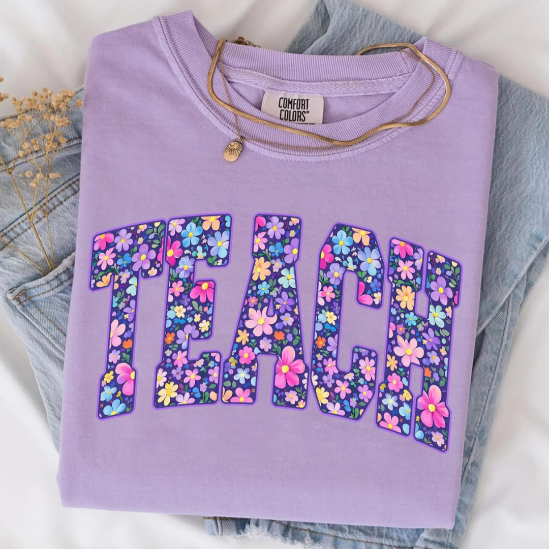 Lavender purple graphic tee featuring oversized floral "TEACH" collegiate lettering in colorful pink, blue, yellow, and purple flowers.
