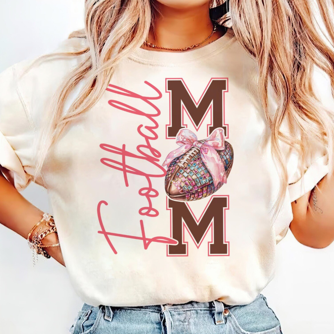 Natural graphic tee featuring a Football Mom design with bold varsity lettering, a pink disco football with a bow, and pink script text.