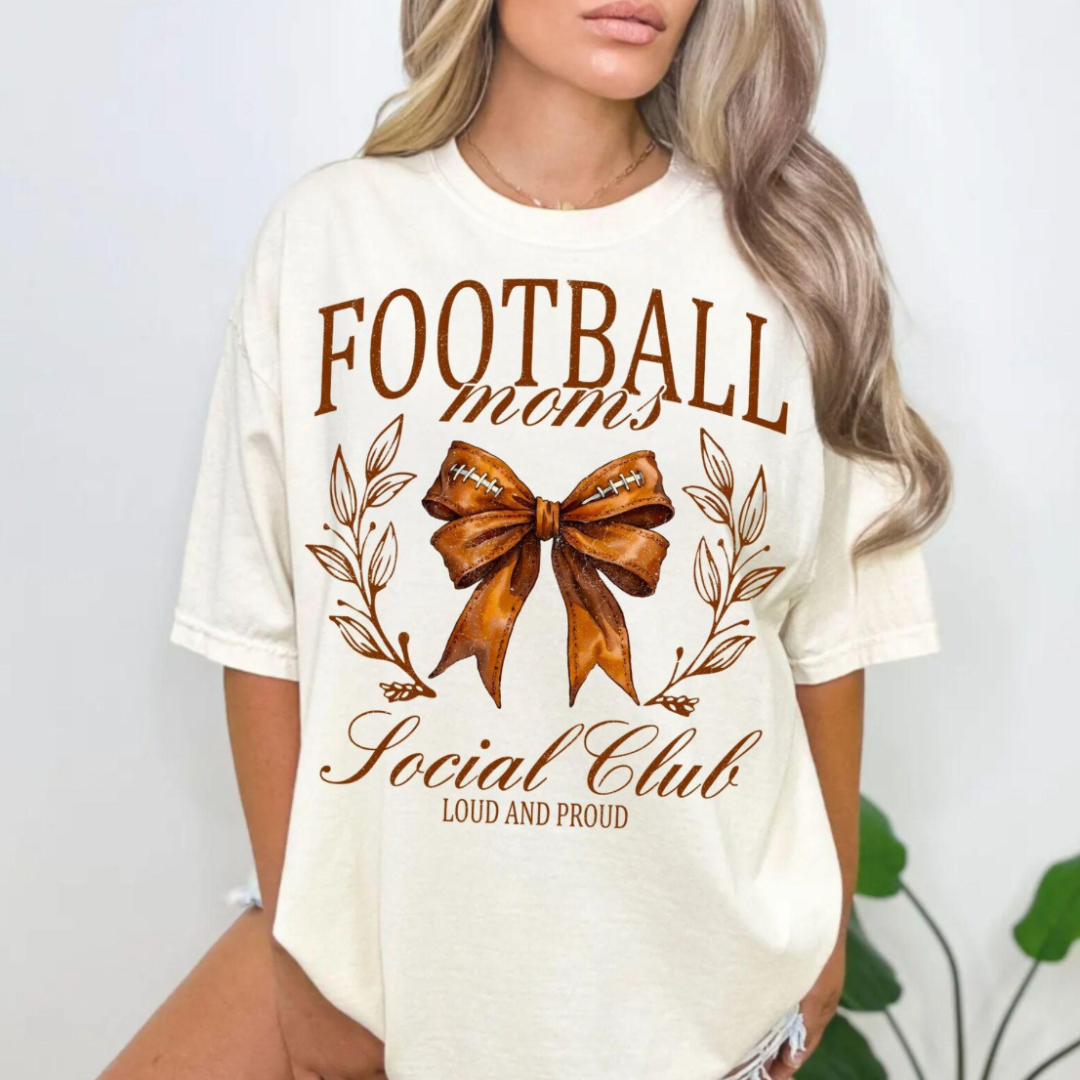 White graphic tee featuring the phrase "Football Moms Social Club – Loud and Proud" with a football-inspired bow and botanical accents.