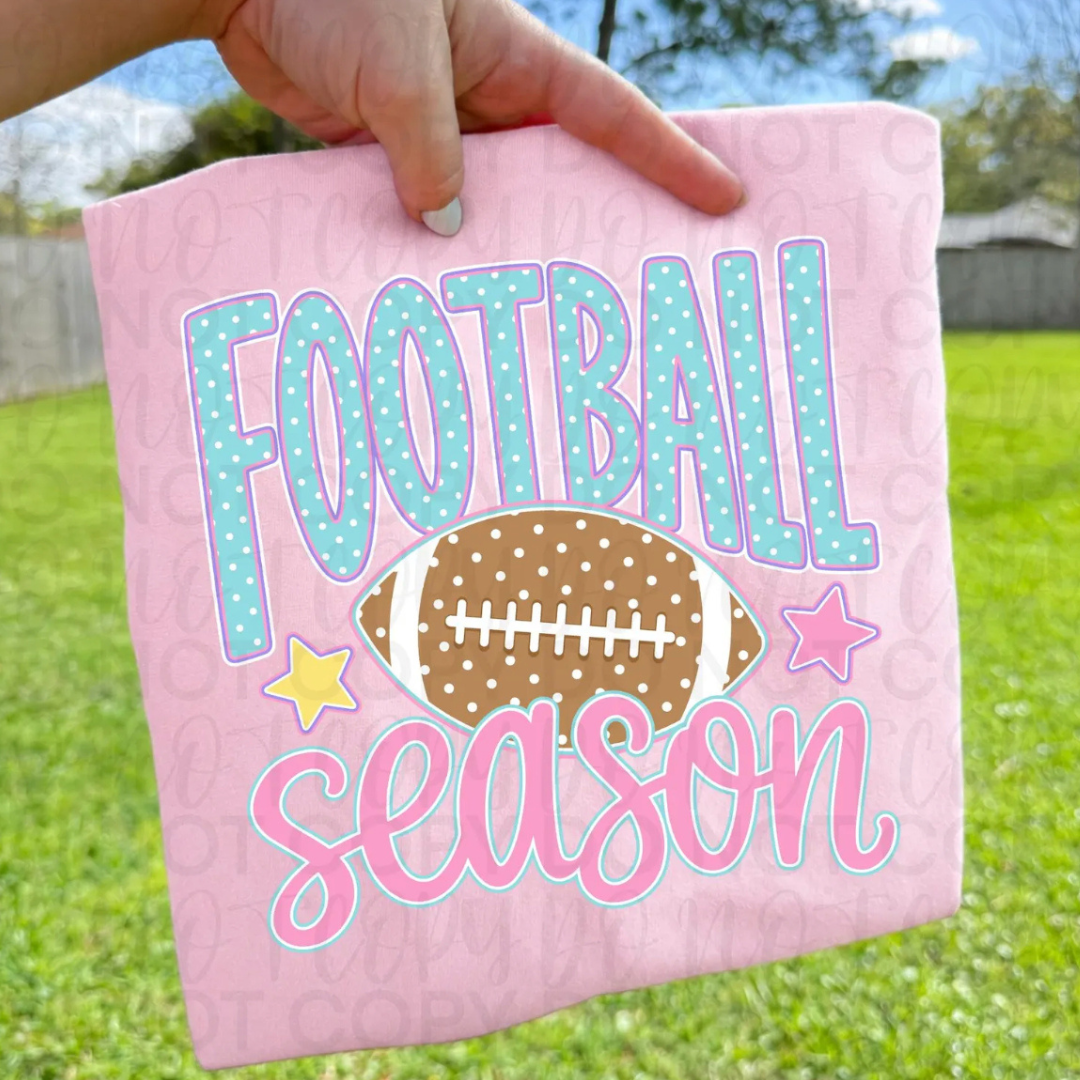 Light pink graphic tee featuring aqua polka dot "Football" lettering, pink "Season" script, a football graphic, and pastel star accents.