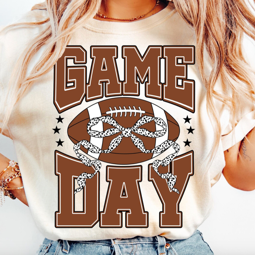 Natural graphic tee featuring bold Game Day lettering with a football and black and white spotted bow, perfect for football season.