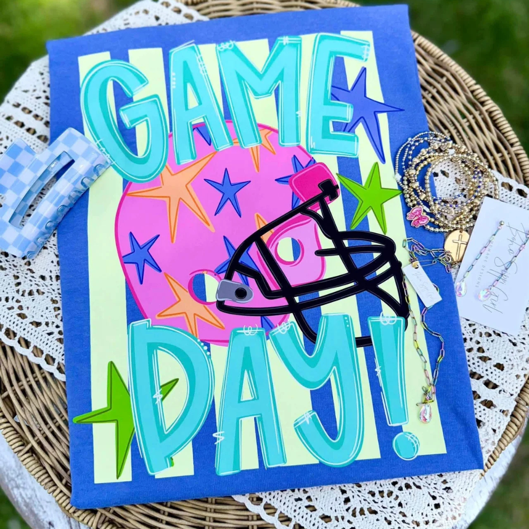 Royal blue graphic tee featuring colorful "Game Day!" lettering with a pink football, black football helmet, stars, and a bright retro-inspired background.