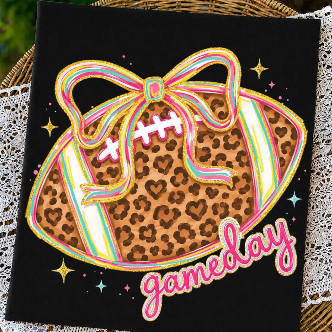 Black graphic tee featuring a leopard print football with a colorful oversized bow and pink "Gameday" lettering.