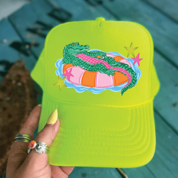 Foam trucker hat available in multiple colors featuring a playful alligator relaxing on a colorful pool float with retro-inspired star accents.