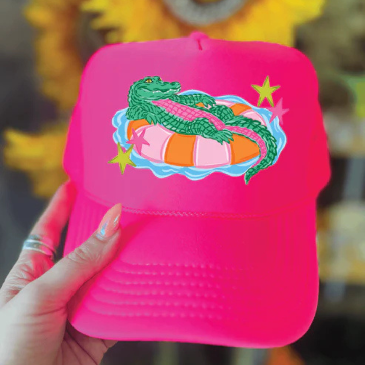 Foam trucker hat available in multiple colors featuring a playful alligator relaxing on a colorful pool float with retro-inspired star accents.
