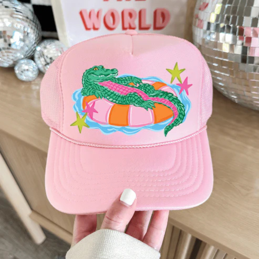 Foam trucker hat available in multiple colors featuring a playful alligator relaxing on a colorful pool float with retro-inspired star accents.