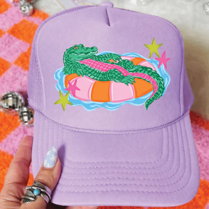 Foam trucker hat available in multiple colors featuring a playful alligator relaxing on a colorful pool float with retro-inspired star accents.