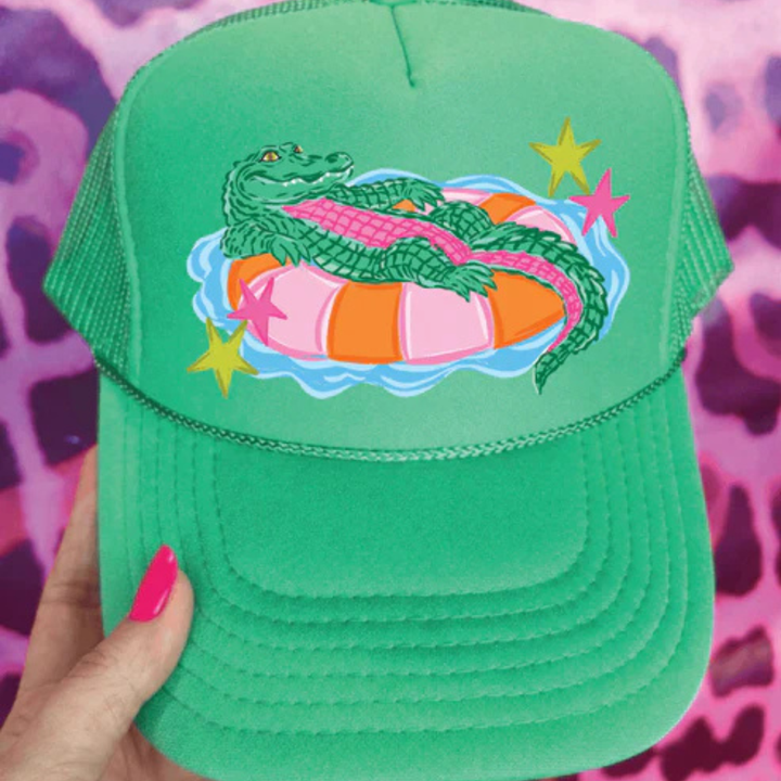 Foam trucker hat available in multiple colors featuring a playful alligator relaxing on a colorful pool float with retro-inspired star accents.