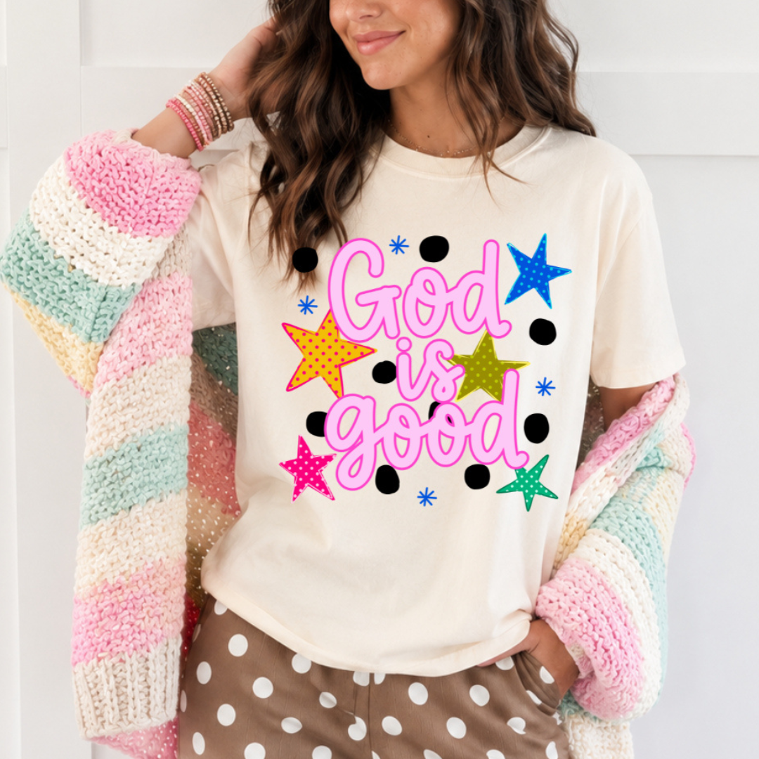 Ivory Christian graphic tee featuring pink God Is Good lettering with colorful stars, black polka dots and bright retro-inspired accents.