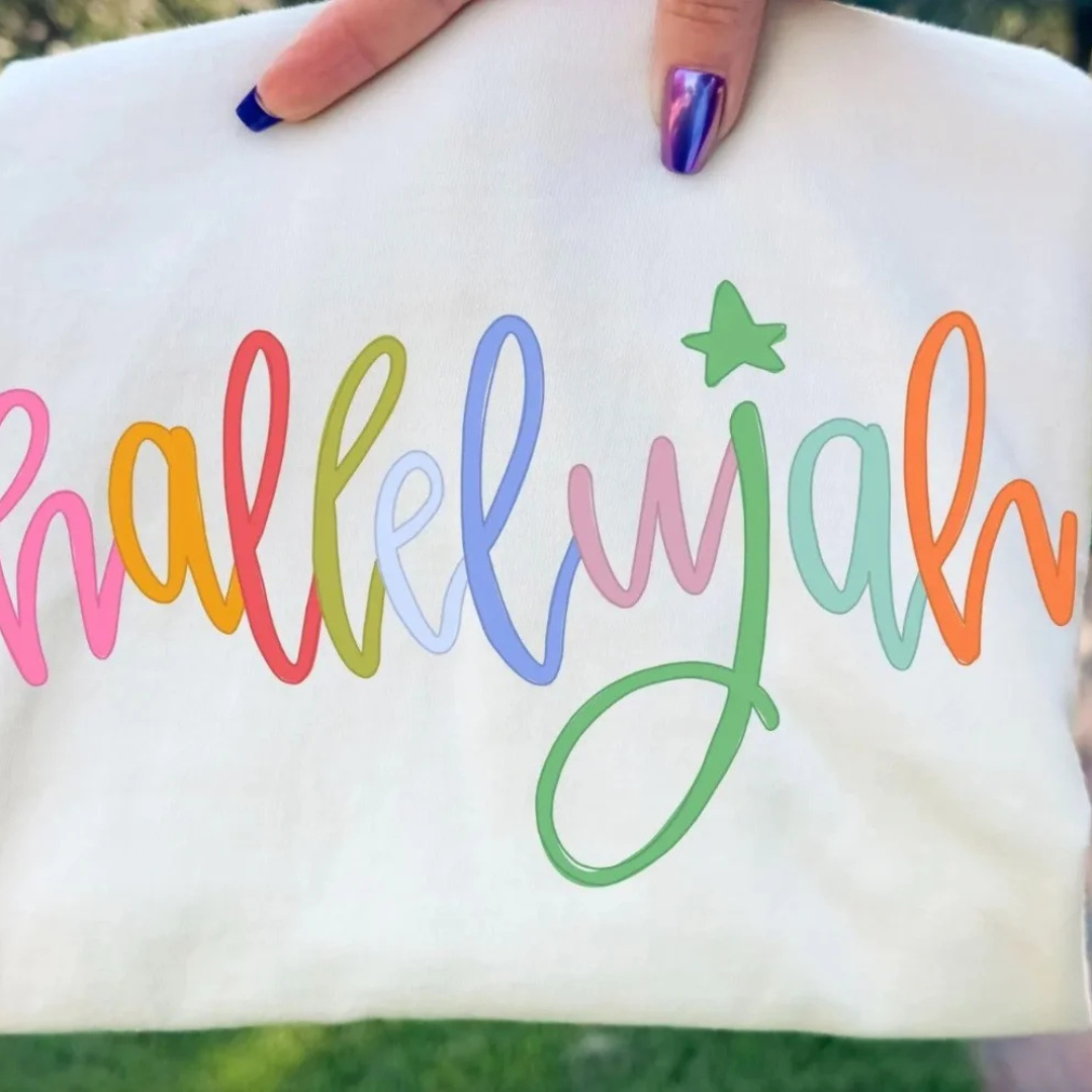 White graphic tee featuring a colorful rainbow script "hallelujah" design with a green star accent.
