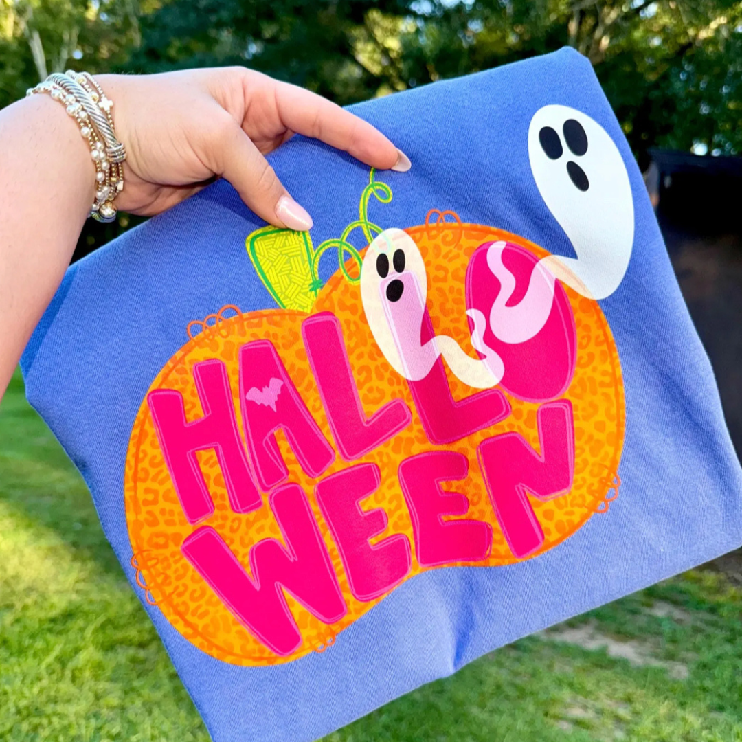 Periwinkle blue Halloween graphic tee featuring bright pink "Halloween" lettering across an orange pumpkin with floating ghosts and colorful spooky accents.