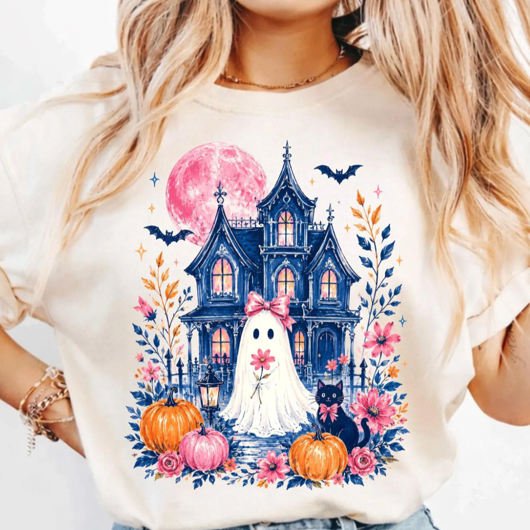 Cream graphic tee featuring a ghost with a pink bow standing in front of a haunted Victorian house with a glowing pink moon, pumpkins, florals, bats, and a black cat.
