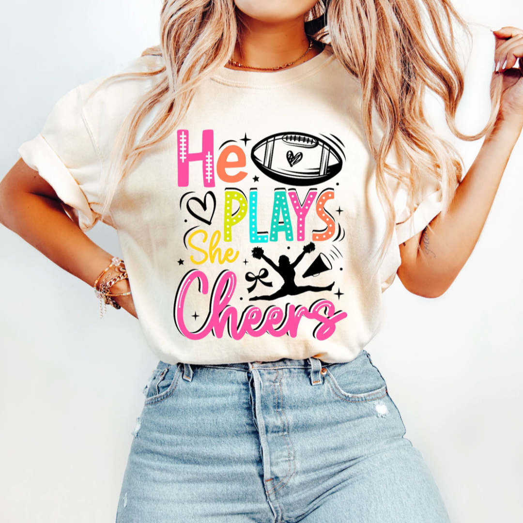 Natural graphic tee featuring the phrase "He Plays She Cheers" with colorful varsity lettering, a football, hearts, and a cheerleader silhouette.
