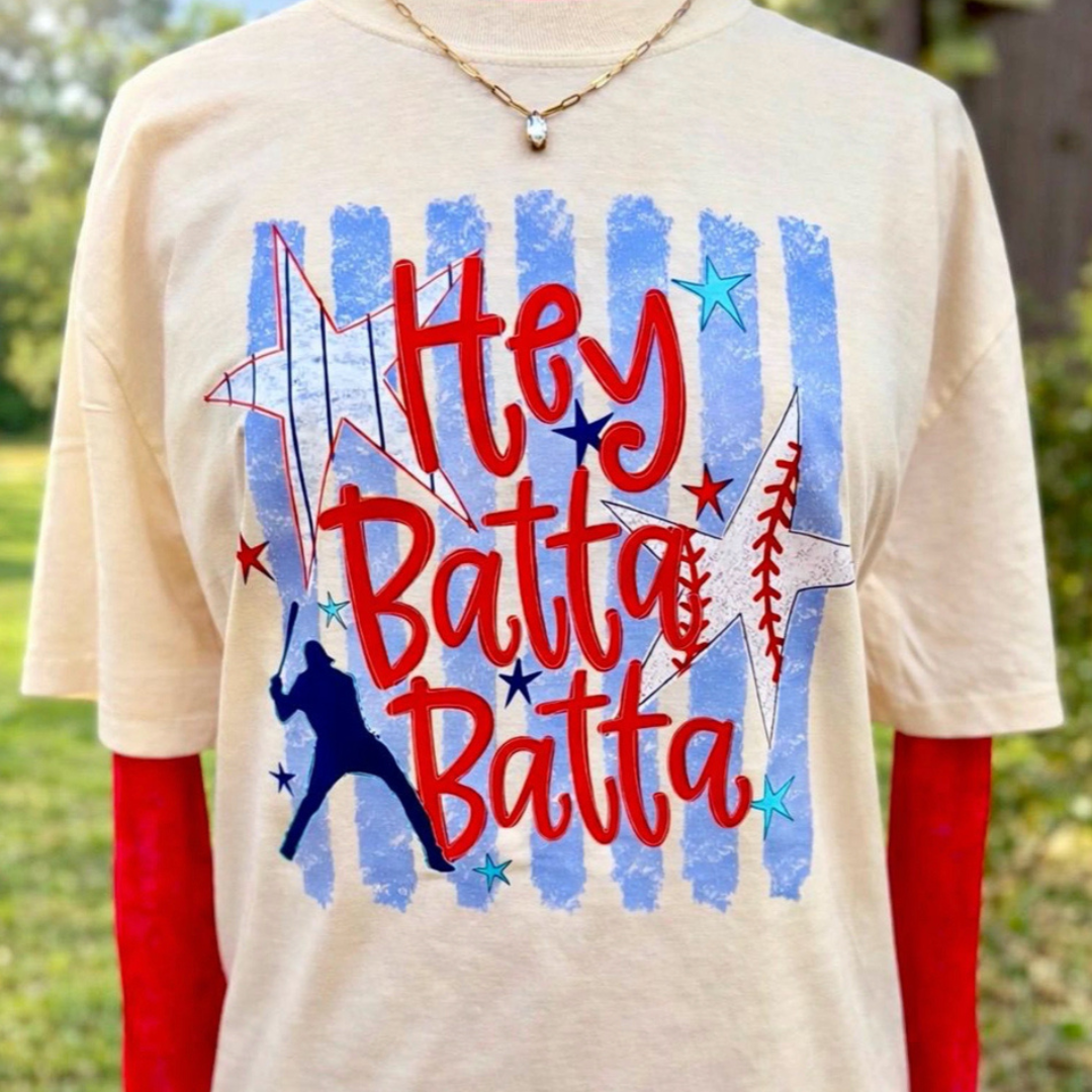 Natural graphic tee featuring the phrase "Hey Batta Batta" with distressed blue stripes, patriotic stars, a baseball batter silhouette, and baseball stitch accents.