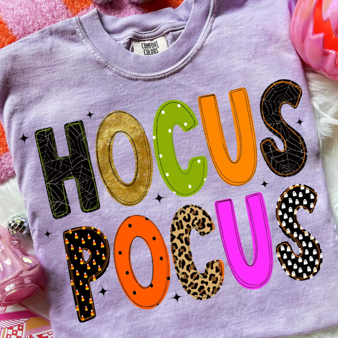Lavender Comfort Colors graphic tee featuring colorful "Hocus Pocus" lettering with Halloween-inspired patterns including spider webs, ghosts, leopard print, candy corn, and polka dots.