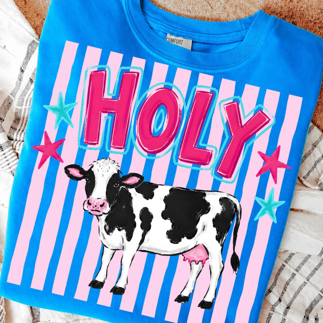 Bright blue graphic tee featuring a black-and-white dairy cow with colorful Holy lettering and pink striped background details.