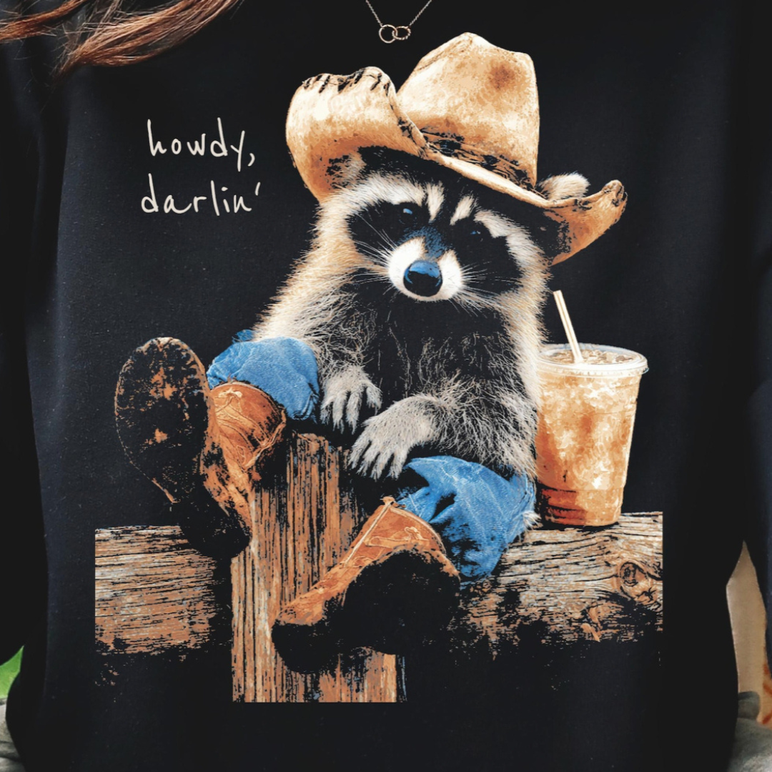 Black graphic tee featuring a raccoon wearing a cowboy hat and boots while holding an iced coffee with the phrase "Howdy, Darlin&