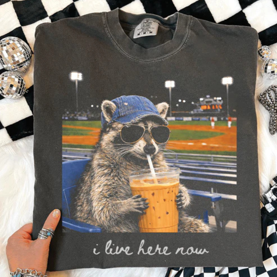 Pepper Comfort Colors t-shirt featuring a funny baseball raccoon wearing sunglasses and drinking iced coffee with the phrase I Live Here Now.