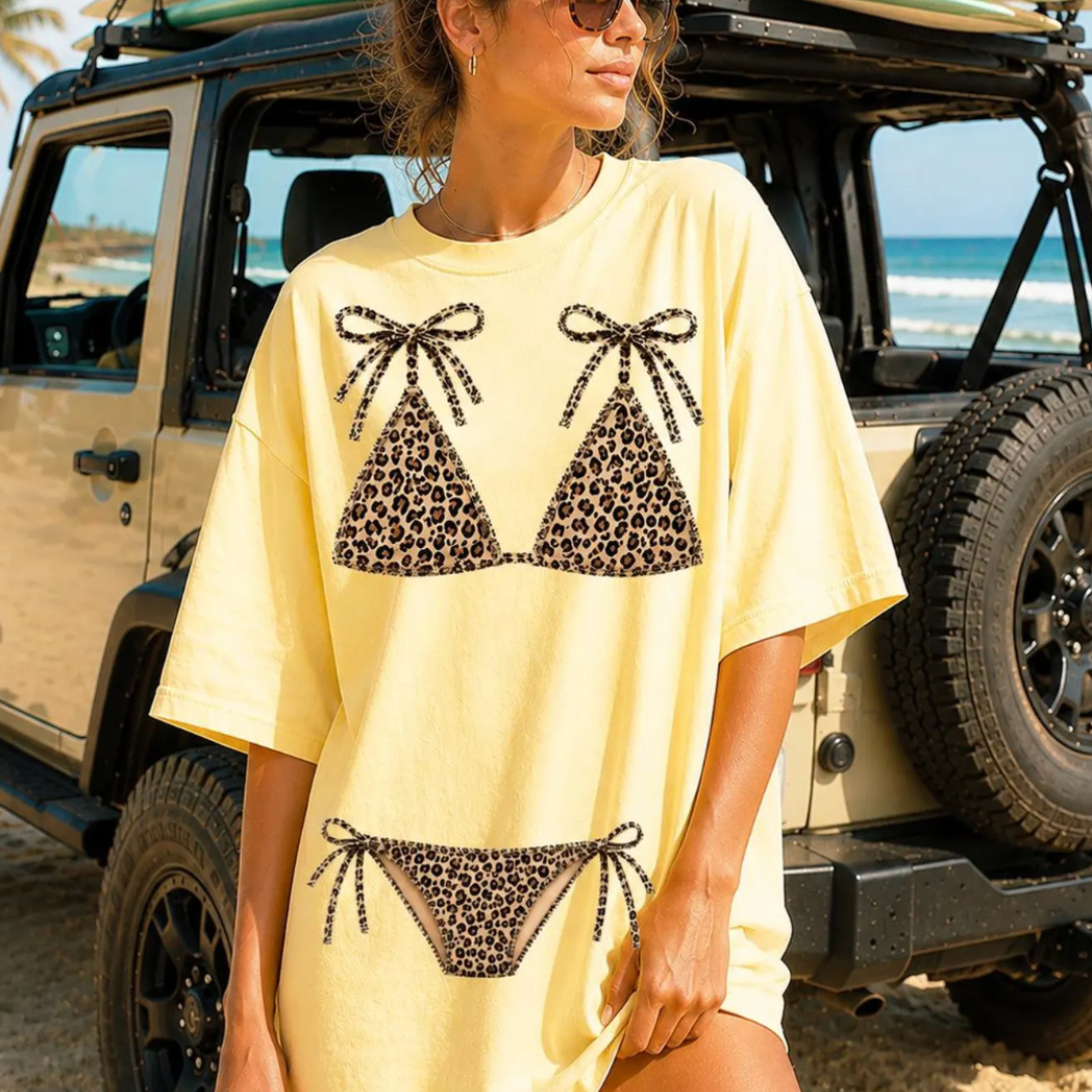 Butter yellow oversized graphic tee featuring a leopard print bikini design with tie details, styled as a fun summer beach and vacation shirt.