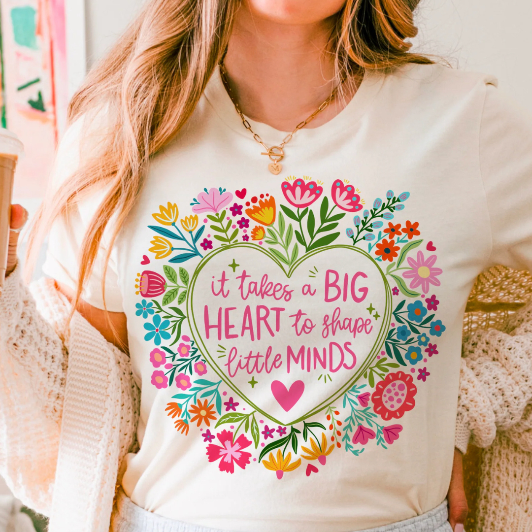 Ivory teacher graphic tee featuring a colorful floral heart with the phrase "It Takes a Big Heart to Shape Little Minds."