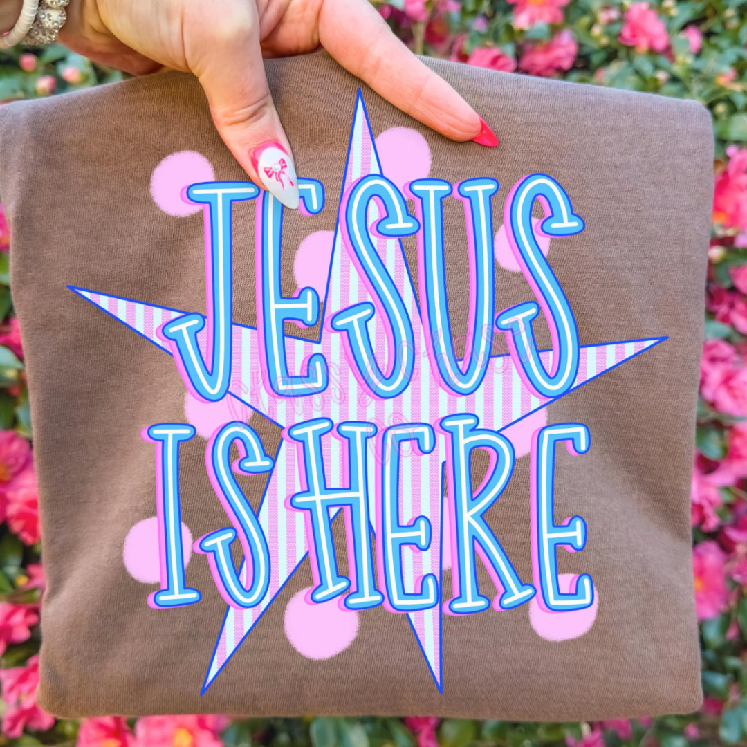 Mocha brown graphic tee featuring a colorful "Jesus Is Here" design with pastel blue lettering, pink accents, and a striped star background.