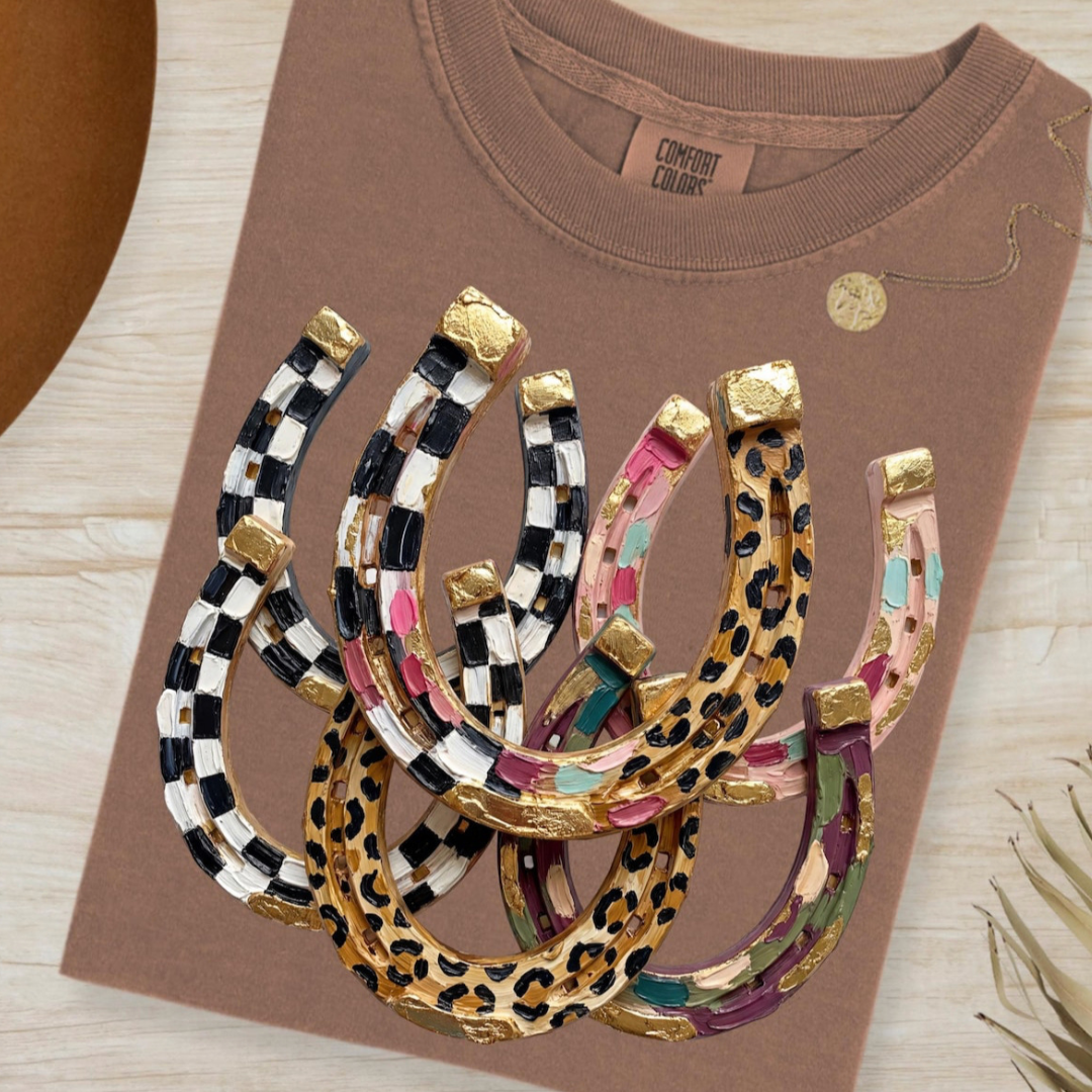 Mauve graphic tee featuring a colorful collection of western horseshoes with checkerboard, leopard, and painted pattern details.