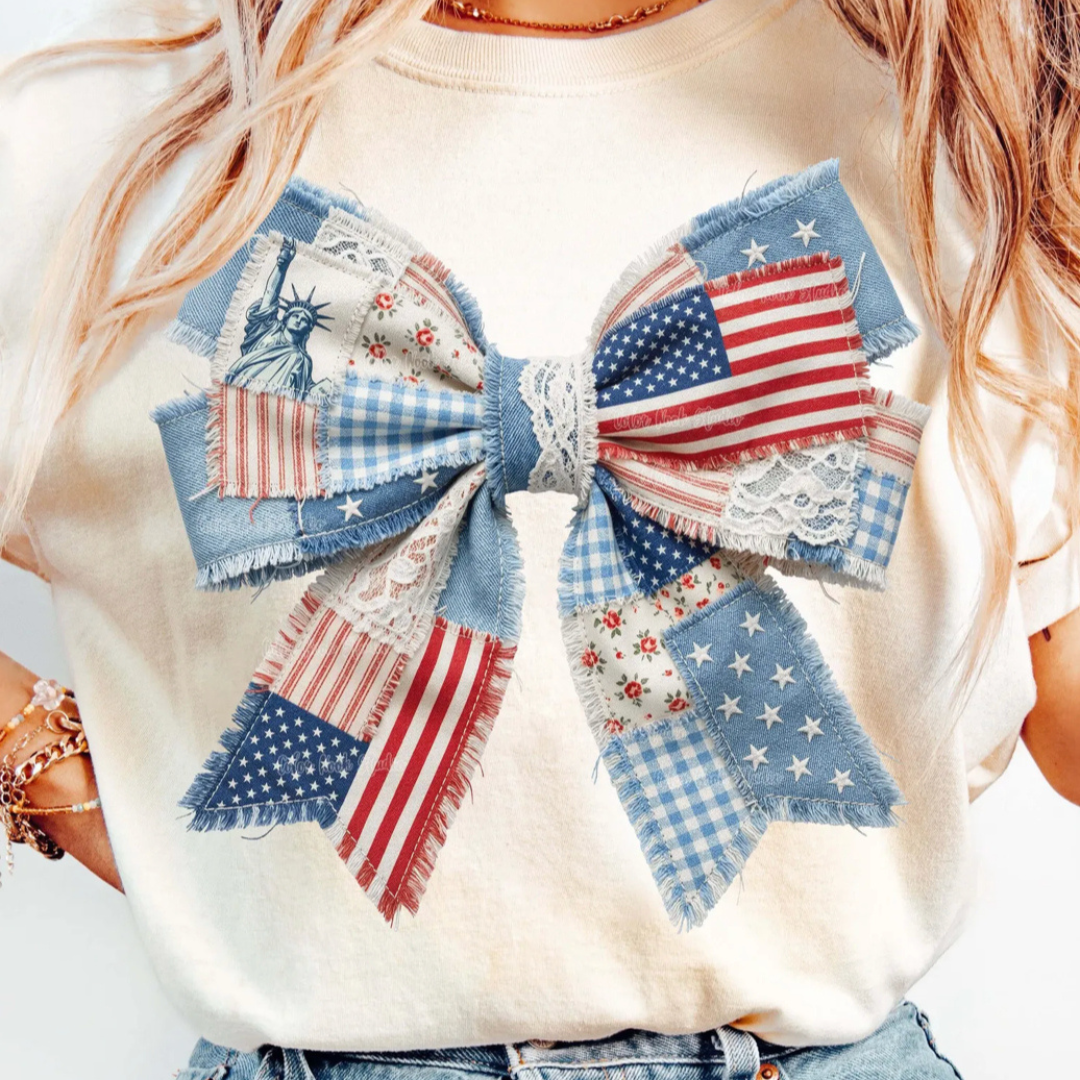Natural cream graphic tee featuring a large patchwork patriotic bow with stars, stripes, gingham, floral prints, denim details, and lace accents.