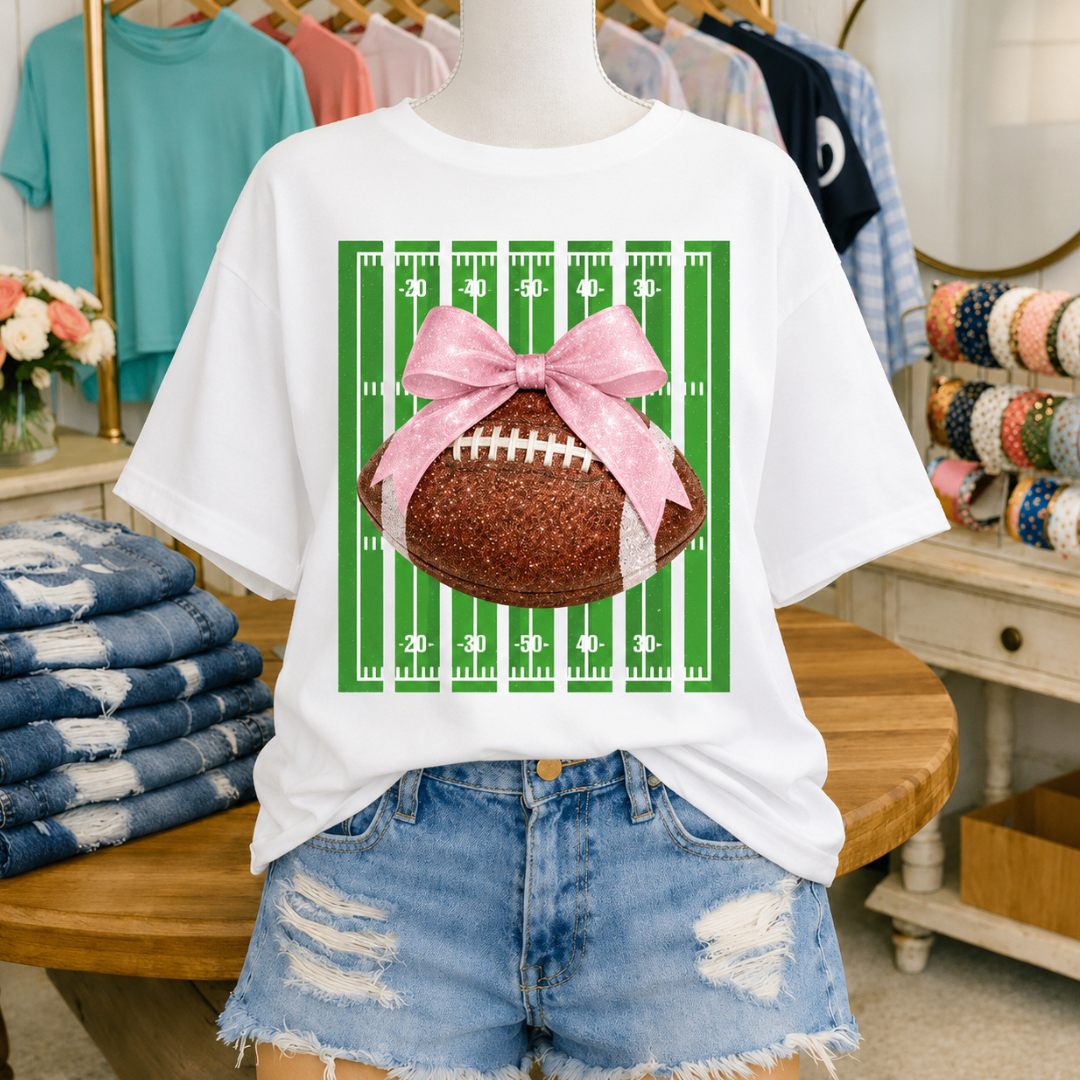 White graphic tee featuring a football field with a glitter-inspired football topped by a pink bow for a feminine football game day look.
