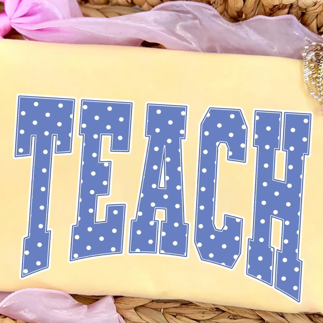Butter yellow graphic tee featuring oversized blue collegiate-style "TEACH" lettering with a white polka dot pattern.