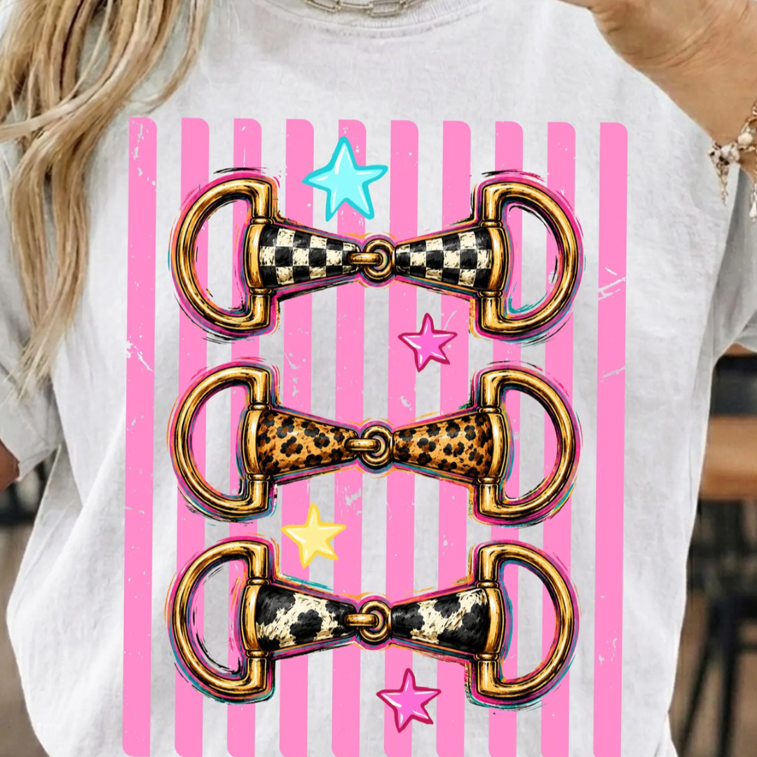 White graphic tee featuring three western snaffle bits with checkered, leopard, and cow print details on a pink striped background.