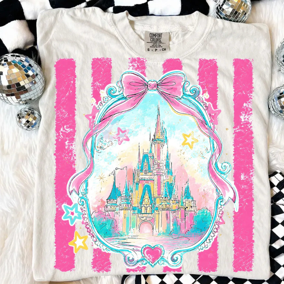 Ivory Comfort Colors graphic tee featuring a pastel fairytale castle inside an ornate frame with a pink bow, stars, hearts, and pink distressed stripe background.