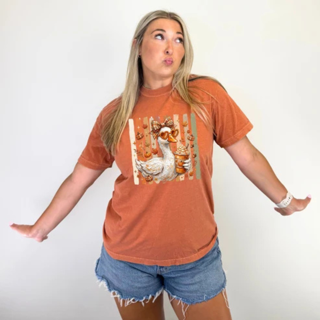 Burnt orange graphic tee featuring a whimsical goose wearing sunglasses with a bow, holding a pumpkin spice latte and surrounded by pumpkins and fall florals.
