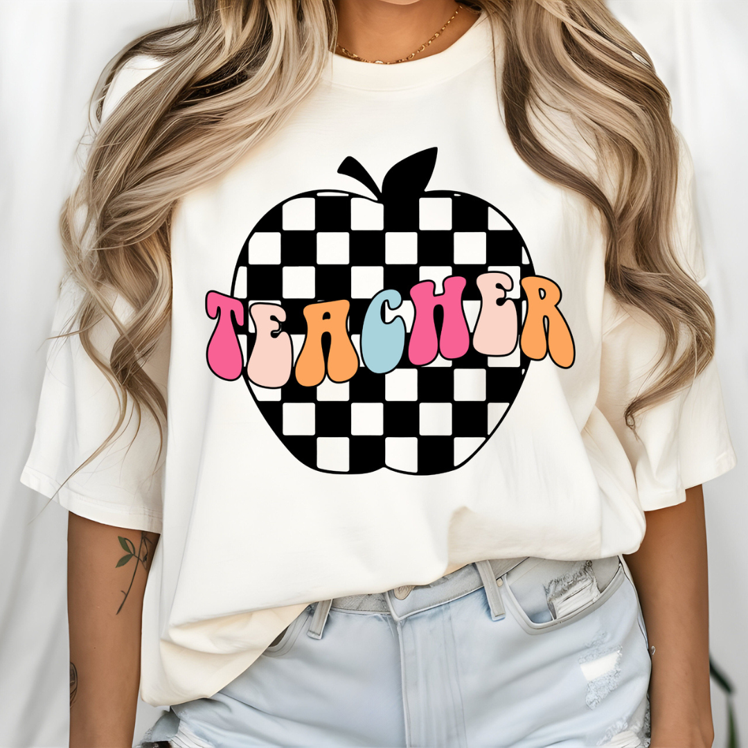 White graphic tee featuring a black-and-white checkerboard apple with colorful retro "Teacher" lettering across the front.
