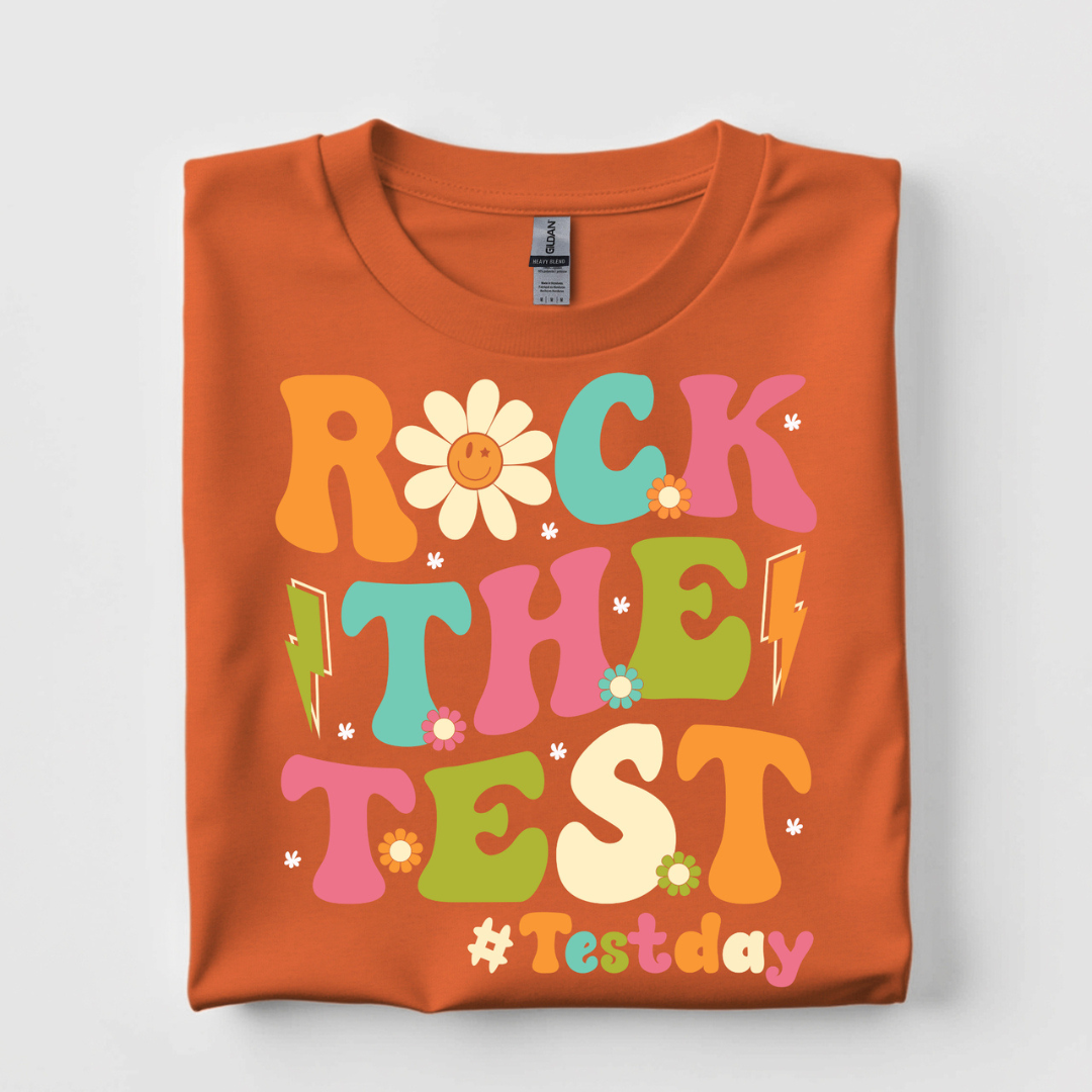 Burnt orange graphic tee featuring colorful retro Rock the Test lettering with flowers, lightning bolts, and a smiley daisy.