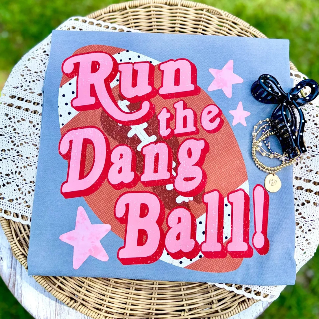 Slate blue graphic tee featuring oversized pink "Run the Dang Ball!" lettering over a football background with pink stars.