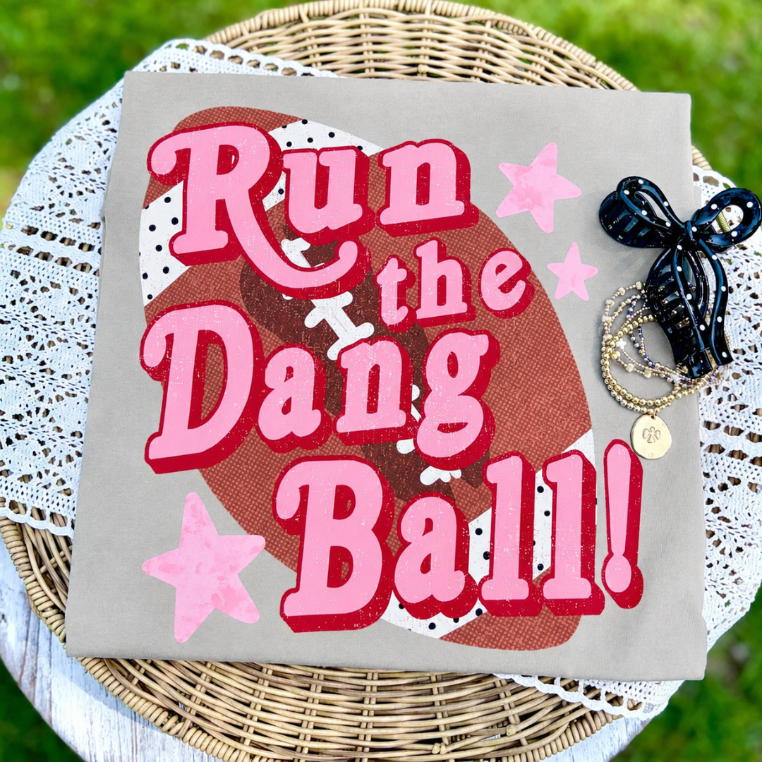 Sand graphic tee featuring oversized pink "Run the Dang Ball!" lettering over a football graphic with pink stars.