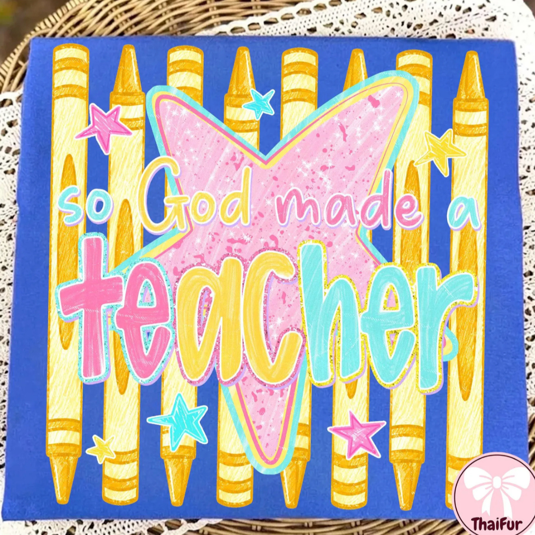 Royal blue teacher graphic tee featuring colorful crayons, a pink cross, stars, and the phrase "So God Made a Teacher."