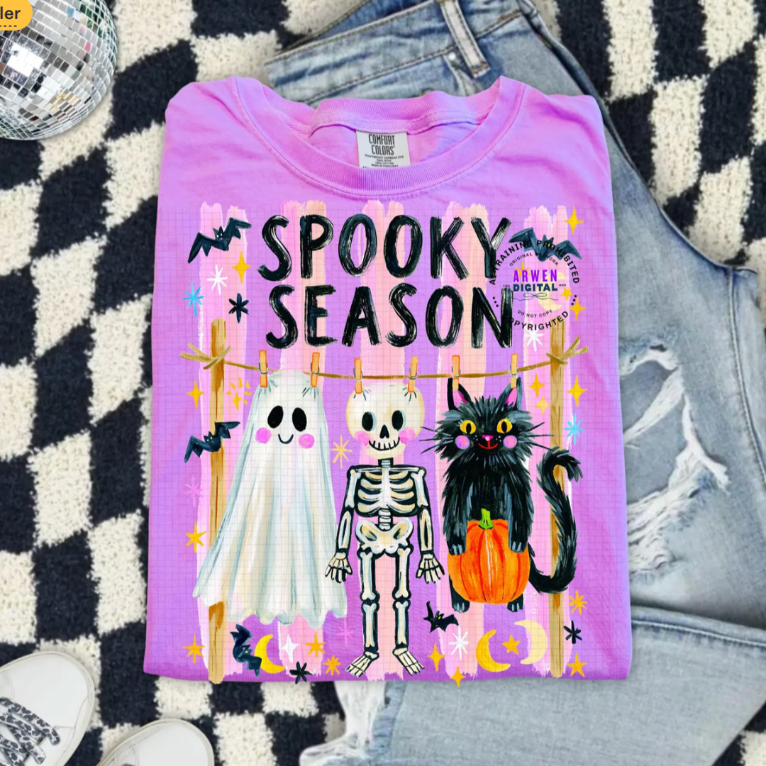 Orchid purple Comfort Colors graphic tee featuring a smiling ghost, skeleton, and black cat hanging on a clothesline with "Spooky Season" lettering and whimsical Halloween accents.