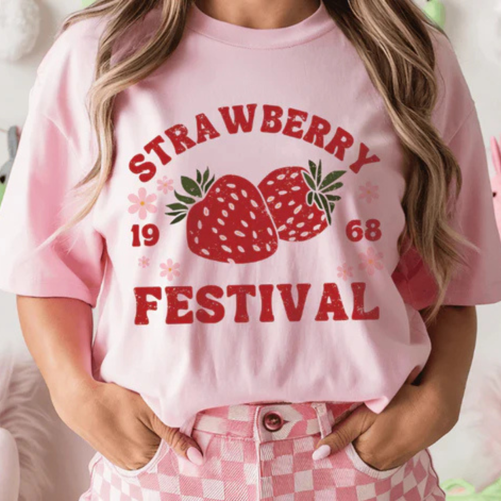 Pink and cream graphic tee featuring a Strawberry Festival design with strawberries, floral accents, and vintage-inspired lettering.