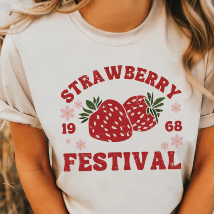 Pink and cream graphic tee featuring a Strawberry Festival design with strawberries, floral accents, and vintage-inspired lettering.