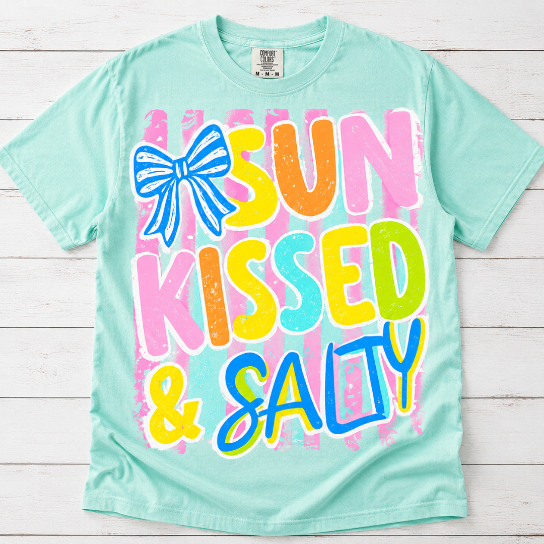 Mint green graphic tee featuring colorful Sun Kissed &amp; Salty lettering, a blue bow accent, and pink striped background.