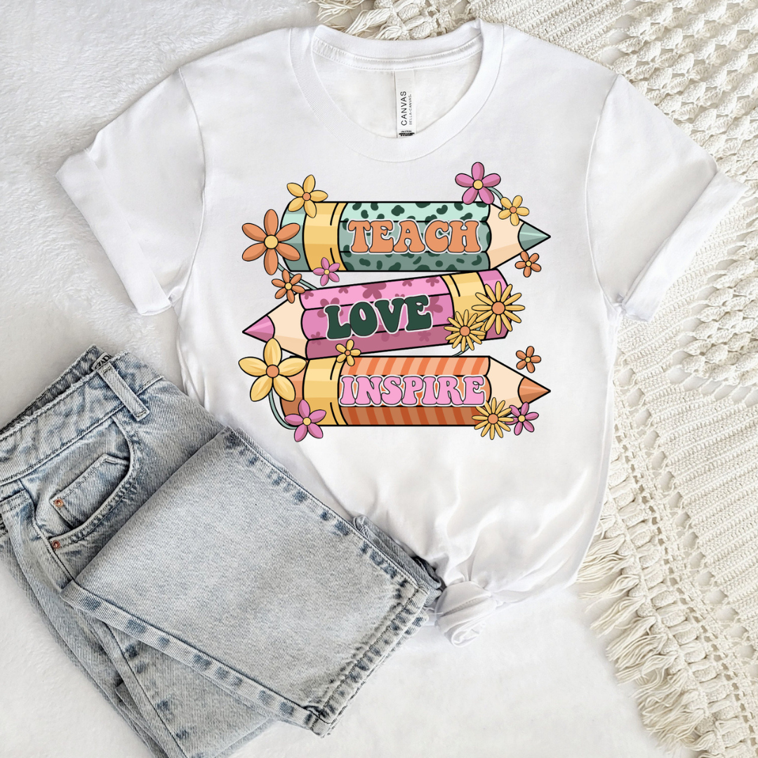 White graphic tee featuring three colorful pencils with the words Teach, Love, and Inspire surrounded by flowers.