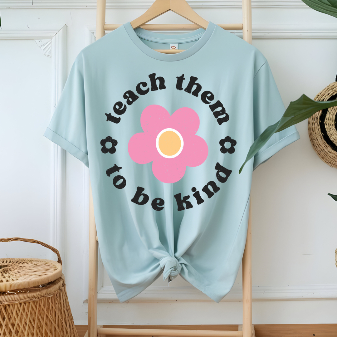 Light blue graphic tee featuring a pink retro flower with the phrase "Teach Them to Be Kind" in black vintage lettering.