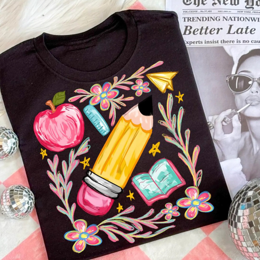 Black graphic tee featuring a colorful oversized pencil surrounded by classroom illustrations including an apple, ruler, flowers, book, and paper airplane.