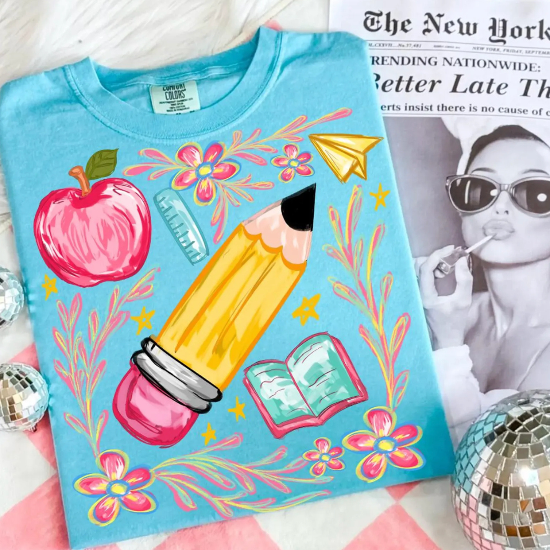 Turquoise blue graphic tee featuring an oversized pencil with colorful classroom illustrations including an apple, ruler, flowers, book, and paper airplane.