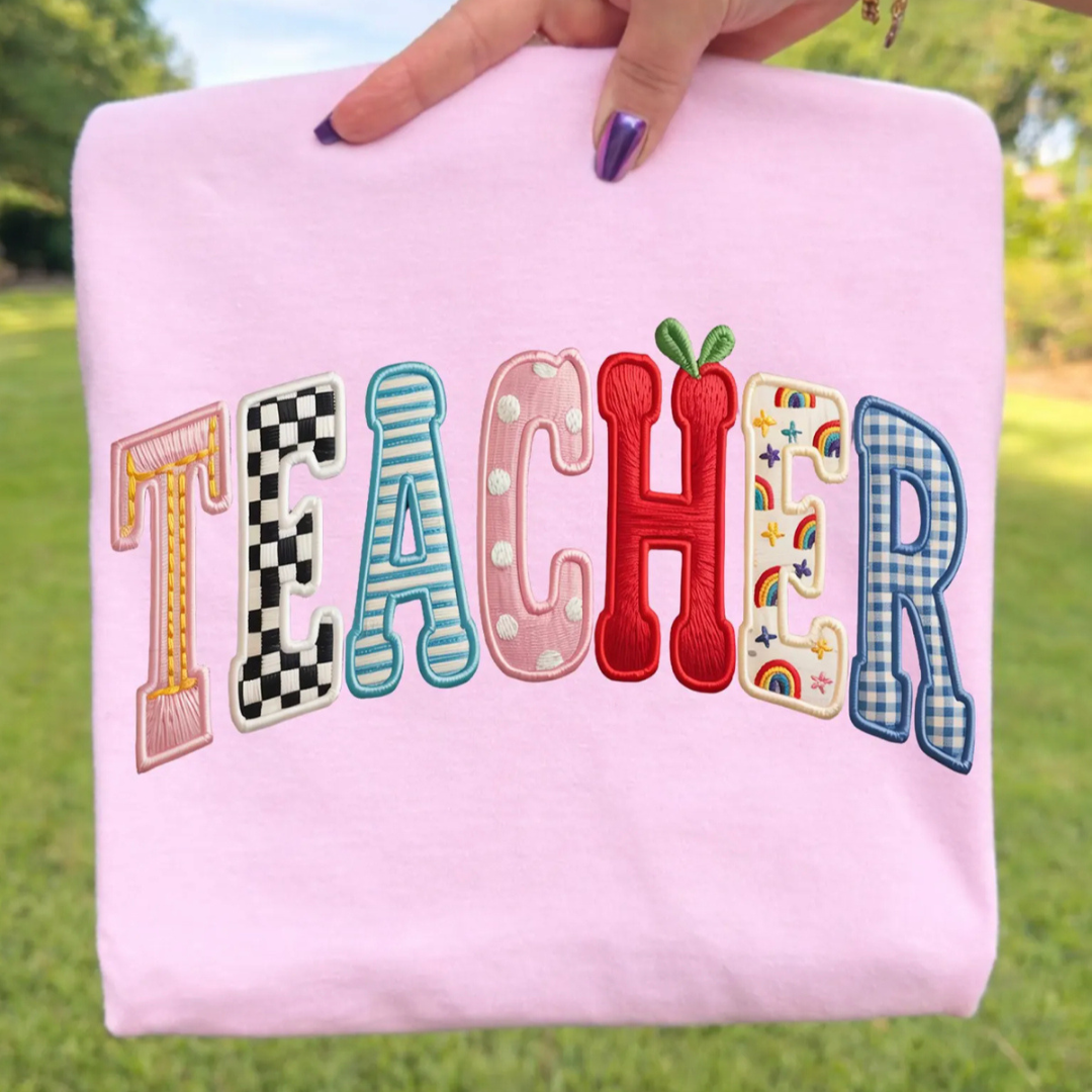 Light pink graphic tee featuring oversized varsity-style "Teacher" lettering with colorful faux embroidered patchwork patterns including checkers, gingham, rainbows, stripes, and an apple.